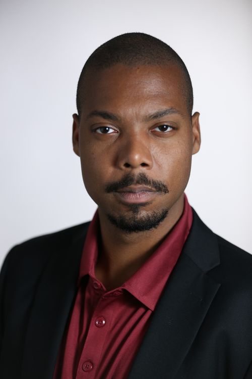 Un homme en veste de costume sombre et chemise, photographié en studio.